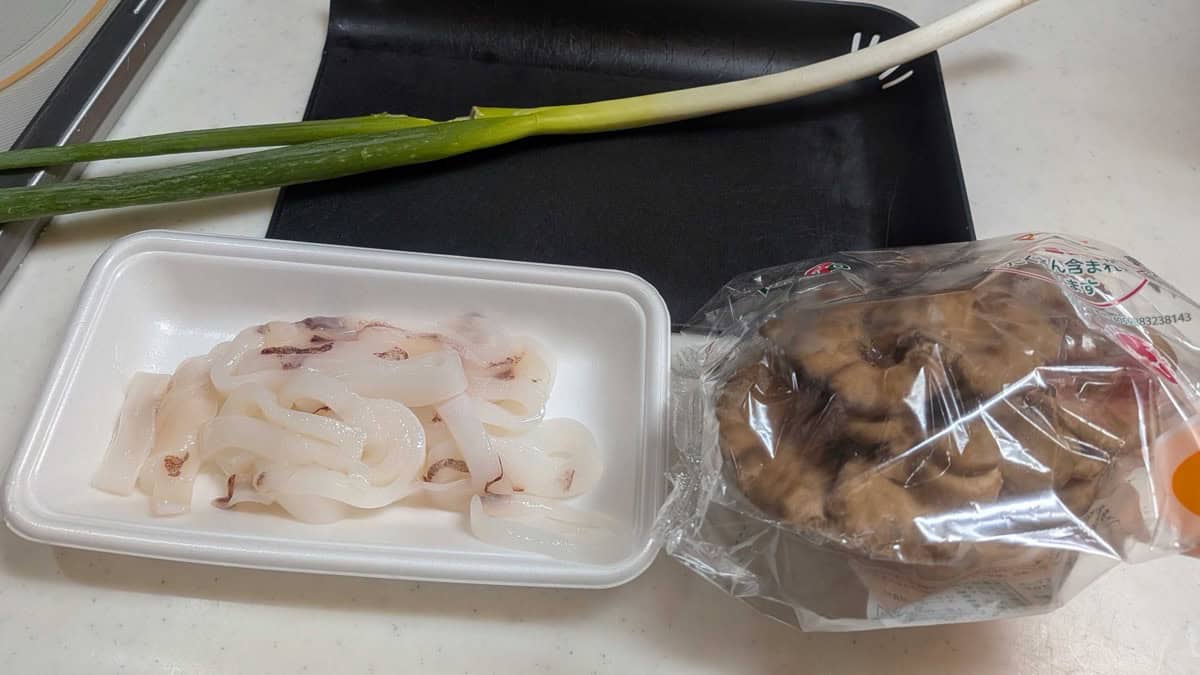 A supermarket tray of cut squid rings and tentacles, a bag of maitake mushrooms, and a whole spring onion on the kitchen counter