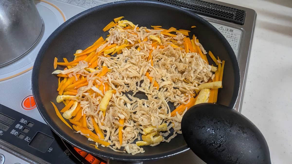 Rehydrated daikon, carrot matchsticks and strips of fried tofu stir-frying in a black pan on an IH cooktop