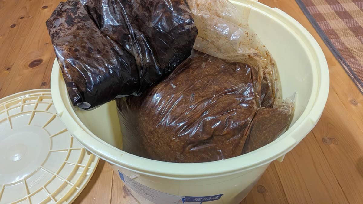 Bags of homemade rice miso of different shades stored in a large plastic bucket on a wooden floor
