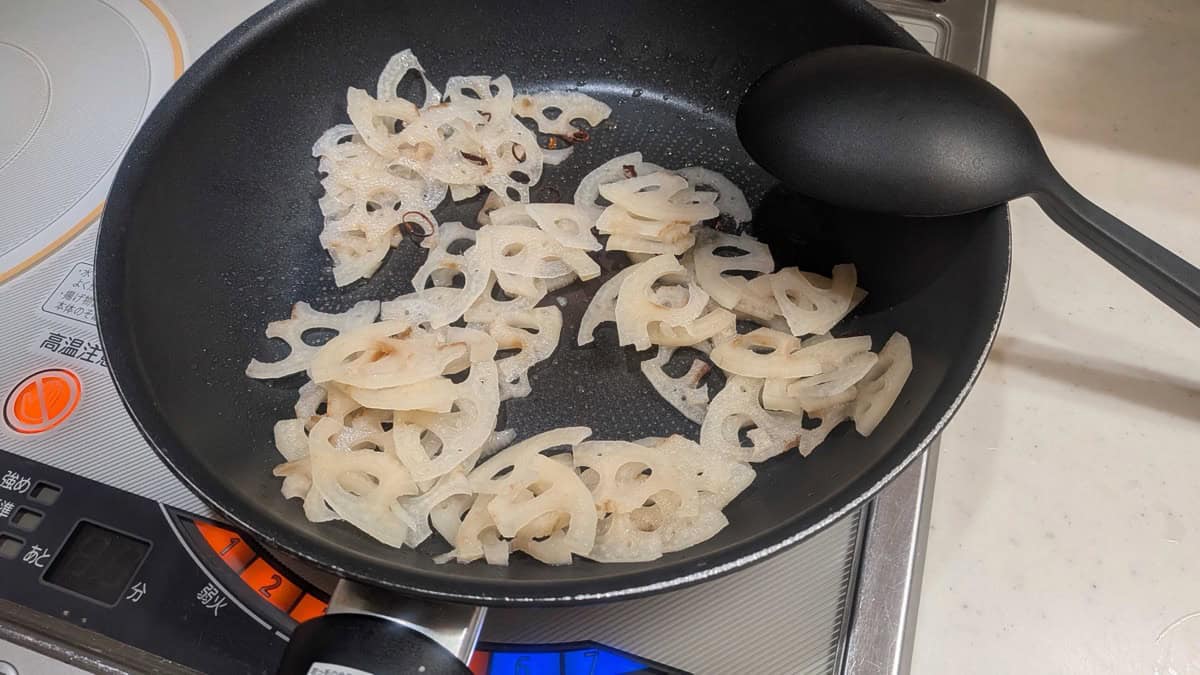 Thin lotus root half-moons stir-frying with dried chili rings in a black frying pan on an IH cooktop