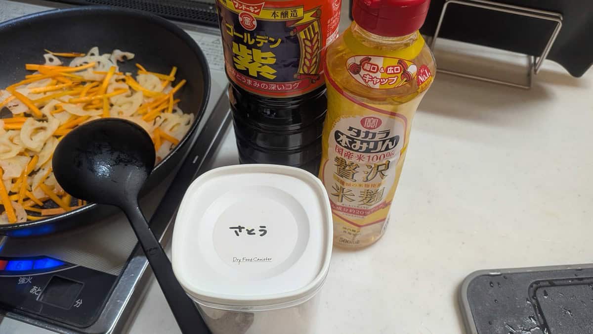 Japanese soy sauce, Takara hon-mirin, and a sugar canister on the kitchen counter next to a pan of lotus root and carrot