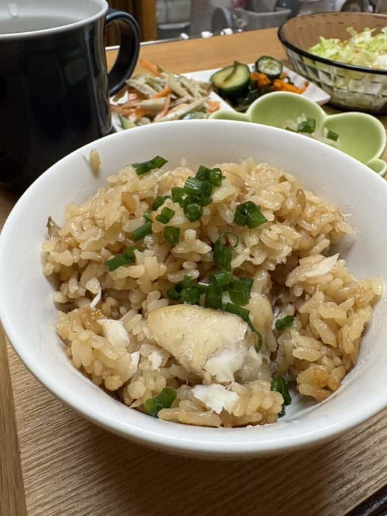 Tai meshi — Japanese sea bream rice with flaked fish and chopped green onion in a white bowl, with side dishes behind