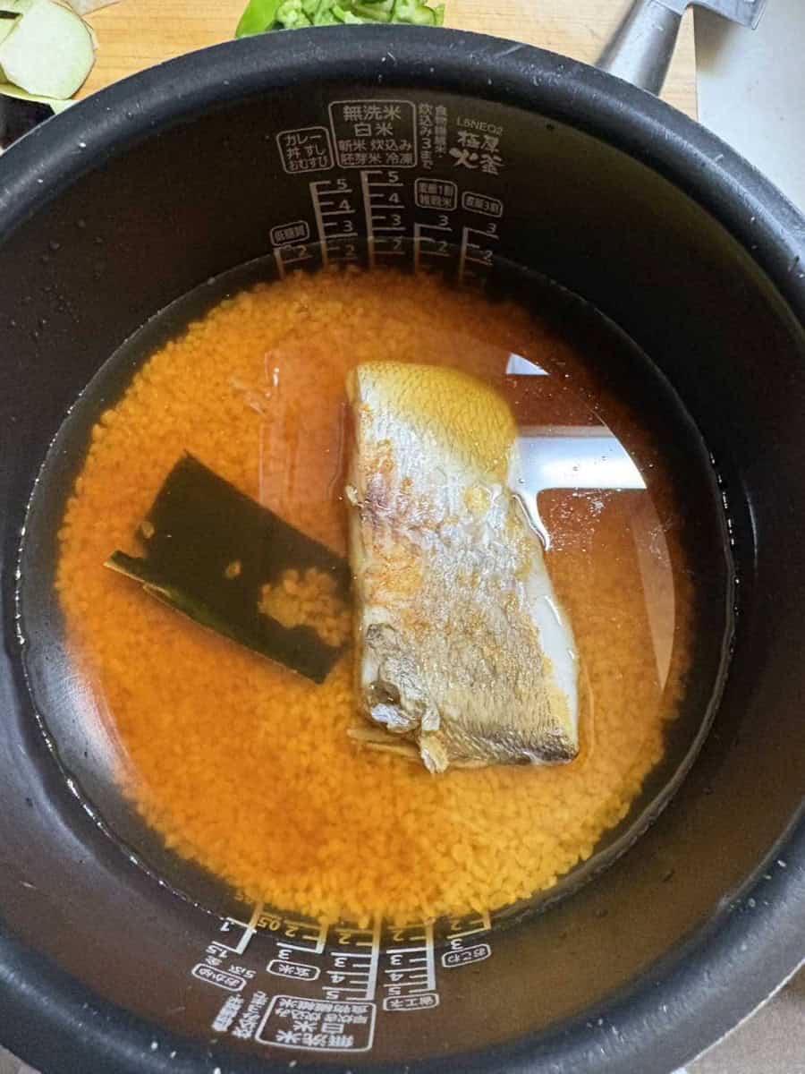 A seared sea bream fillet and kombu sitting on seasoned rice in soy-colored water in a rice cooker pot, ready to cook
