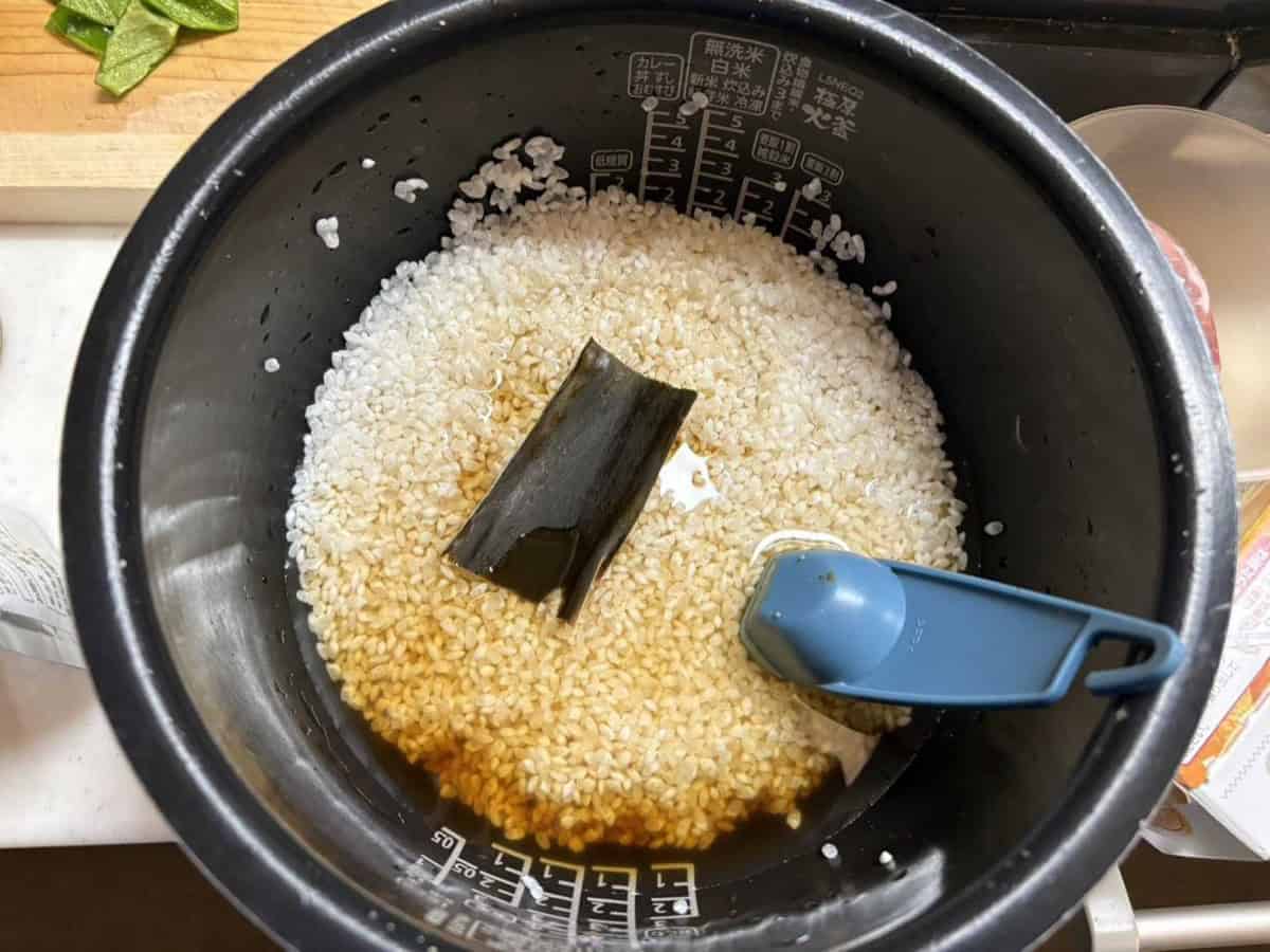 Rinsed rice in a rice cooker pot with soy sauce, sake and mirin poured in and a piece of kombu on top