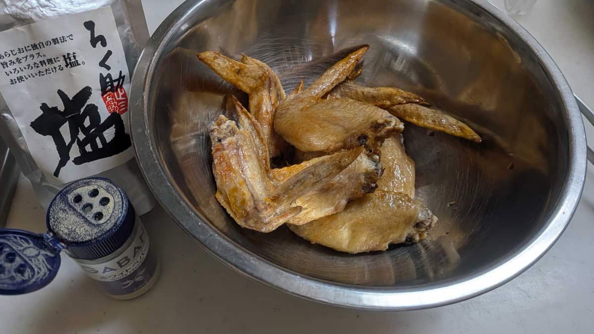 A bag of Japanese Rokusuke umami salt and a white pepper shaker on the counter next to a metal bowl of cooked chicken wings