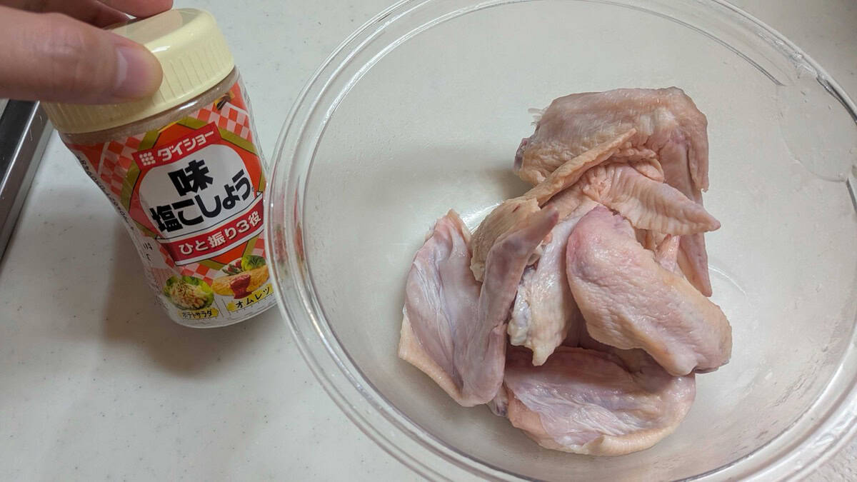 Raw chicken wings in a glass bowl being seasoned from a shaker of Japanese seasoned salt and pepper