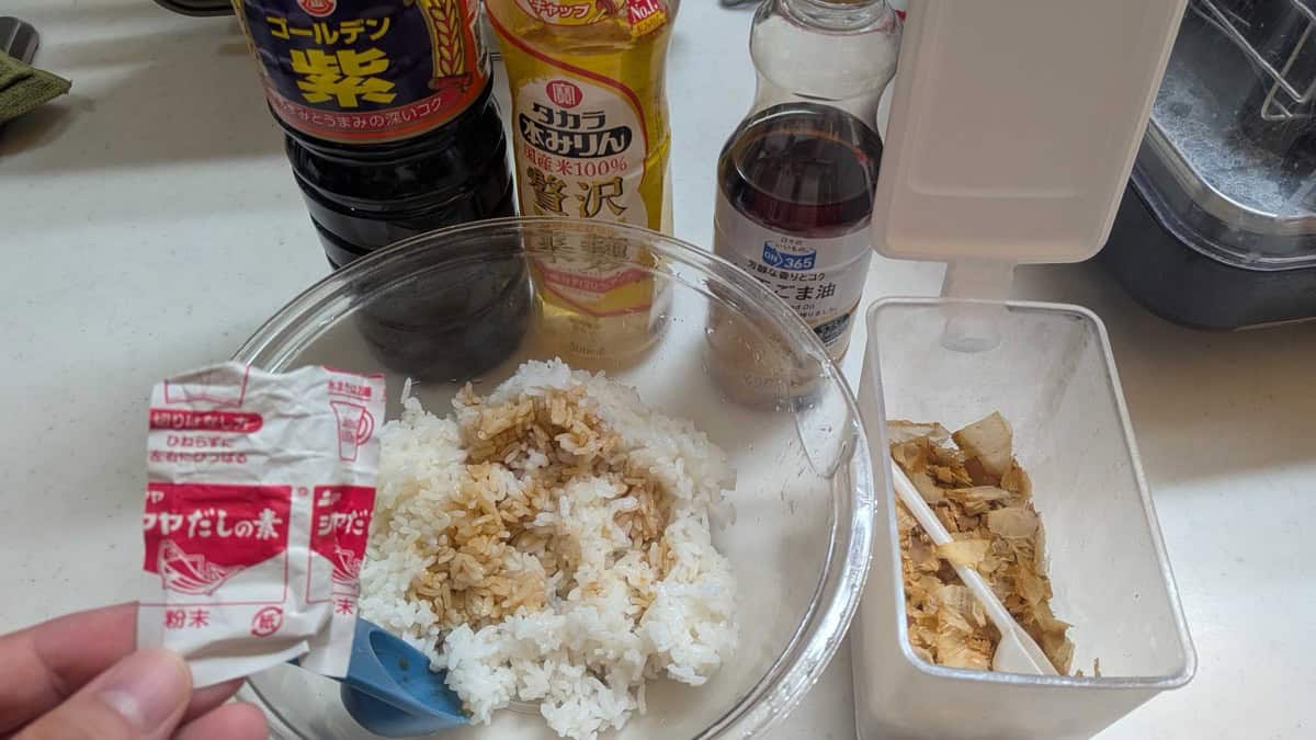 Soy sauce, hon-mirin, sesame oil, a sachet of dashi powder and a container of katsuobushi bonito flakes on a kitchen counter next to a bowl of rice